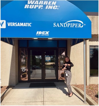 A College of Business student, Makenna Heimlich, standing in front of the company sign, Warren Rupp Inc. creating the Z sign with her hands.