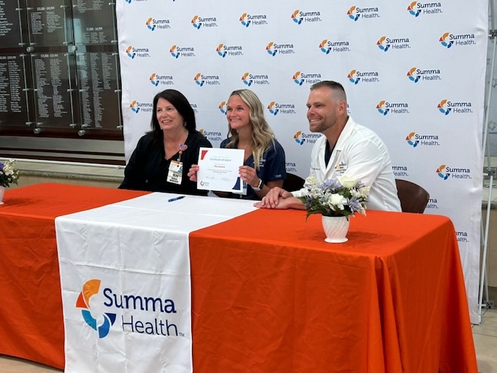 Three people sit at a table with a Summa Health logo. The woman in the middle holds a certificate and smiles, while the others smile at the camera. The table is covered with an orange and white cloth and small flower vases.