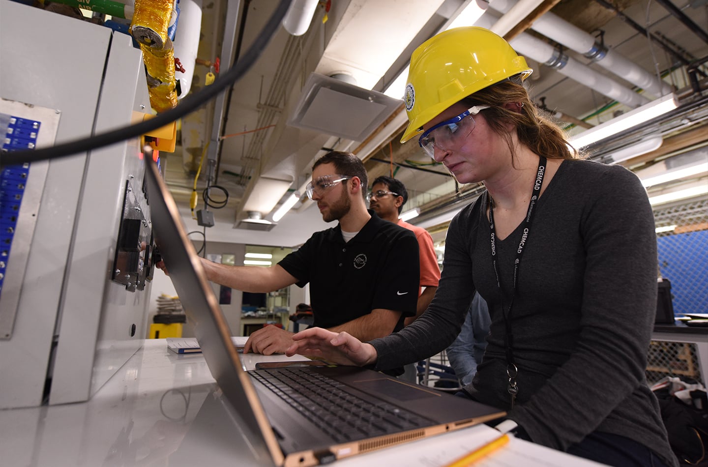 Researchers wearing safety goggles and a hard hat work at a laptop computer inside an industrial laboratory facility, with a third team member visible in the background.