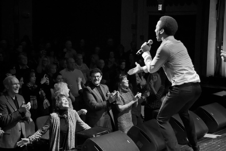 A singer performs energetically on stage as a cheering crowd stands close, clapping and smiling. The scene is lively and captured in black and white.