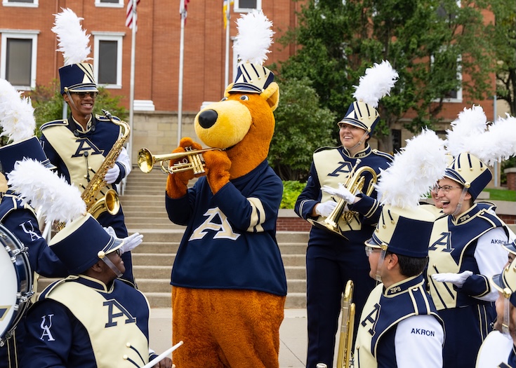 A person in a kangaroo mascot costume plays a trumpet among smiling marching band members in blue and gold uniforms, outdoors near a brick building.
