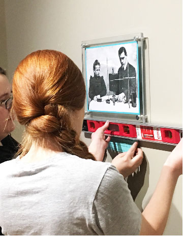 Student carefully aligns a historic photograph on a museum wall using a level during an archival installation.