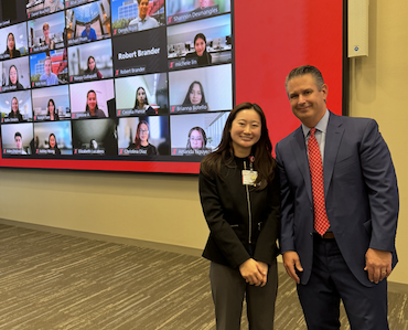 Two people stand and smile inside a conference room; behind them is a large screen displaying a virtual meeting with many participants in individual video call windows.
