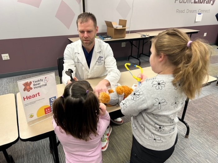 A man in a lab coat shows two young girls how to check a stuffed animal’s heart at a library event. A sign reads “Teddy Bear Hospital: Heart.” Medical toys and boxes are on the table.