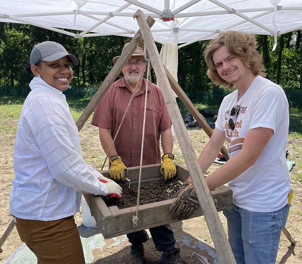 University of Akron archaeology field school students screen soil for artifacts alongside an instructor at an outdoor dig site.