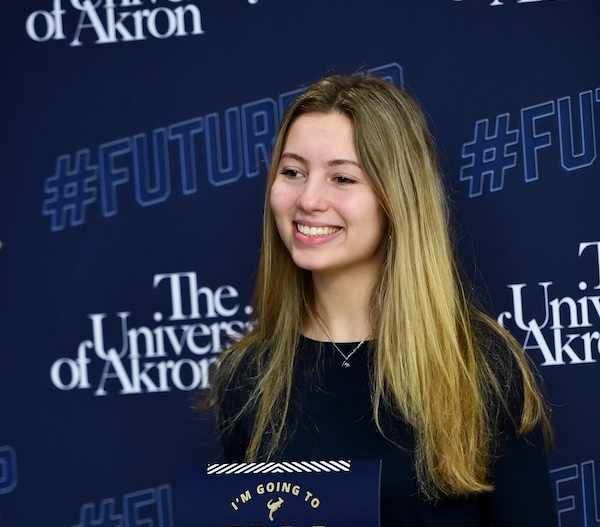 A young woman with long blonde hair smiles in front of a University of Akron backdrop, holding a sign that partially reads, IM GOING TO. The background features #FUTURE and university branding.