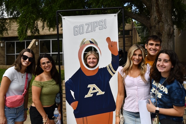 A group of five smiling young adults pose next to a Go Zips! cutout, with one person’s face showing through the cutout. They are outdoors, with trees and a building in the background.