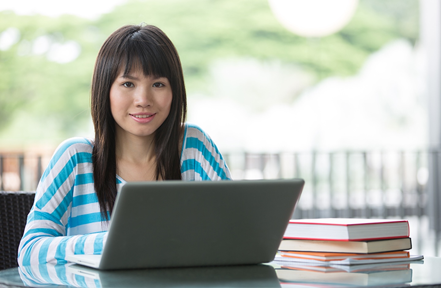 A young woman sits at a glass table outdoors, smiling at the camera. She has a laptop open in front of her and a stack of textbooks beside it. She is wearing a blue and white striped cardigan. Trees and a railing are visible in the blurred background.