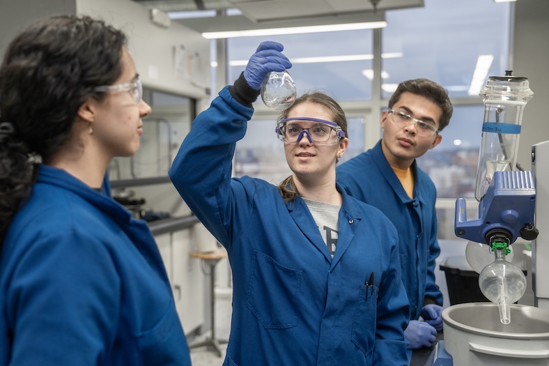 Three people in blue lab coats and safety goggles work in a laboratory; one person examines a flask while the others observe. Lab equipment and windows are visible in the background.