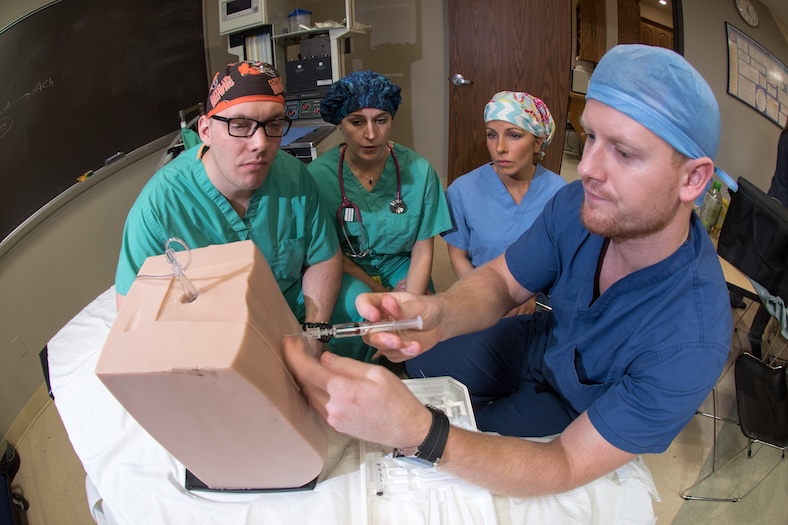 Four medical professionals in scrubs observe as one person demonstrates a procedure with a syringe on a medical training model in a classroom setting.