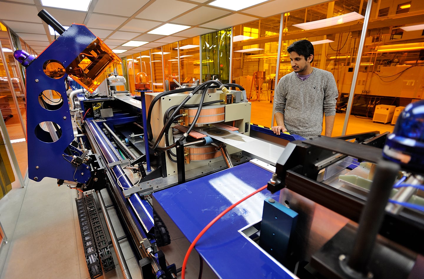 A student observing a large, industrial polymer processing machine in a bright laboratory. The complex equipment features blue supports and copper-coiled components, while large yellow-tinted glass partitions separate the workspace from other areas of the facility.