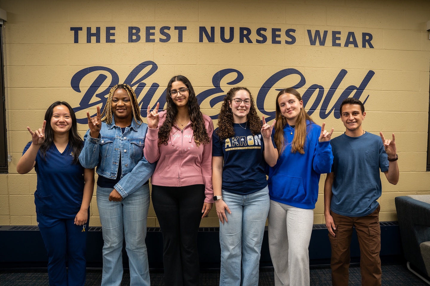Six students stand smiling in front of a wall that says “The Best Nurses Wear Blue & Gold,” making hand gestures. They are dressed in blue and casual clothing.