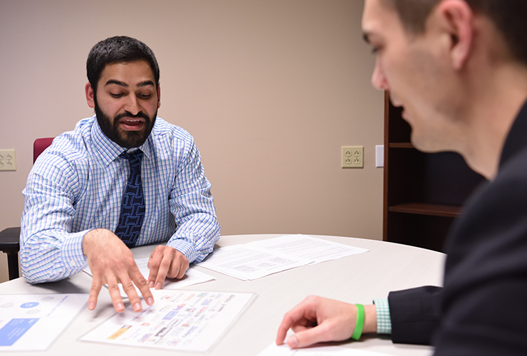 Two men in business attire sit at a table discussing documents; one man gestures to a page with charts while the other listens attentively.