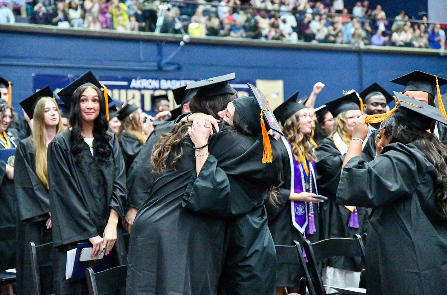 University of Akron graduates in black caps and gowns hug and celebrate on the arena floor during commencement, with other students and spectators in the background.