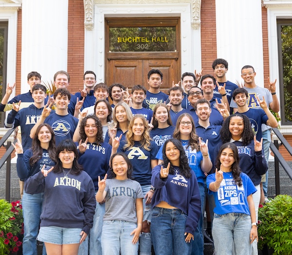 A group of college students in Akron Zips apparel stand on steps in front of Buchtel Hall, smiling and making the Zips hand sign.