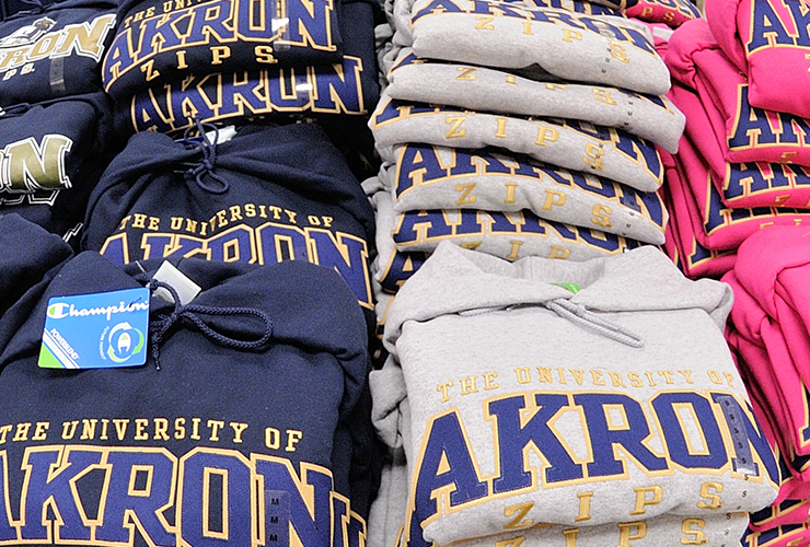 Sweatshirts in navy, gray, and pink, featuring The University of Akron Zips are stacked on a display table. A Champion brand tag is visible on one of the navy sweatshirts.