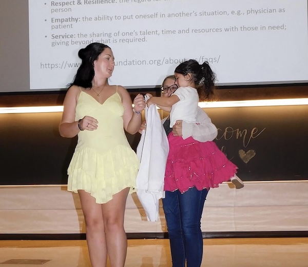 Three people stand on a stage: one woman in a light yellow dress, another adult in jeans and glasses, and a young girl in a pink dress holding a white shirt. A presentation slide is projected behind them.