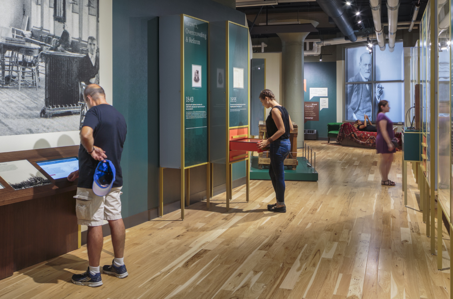 Two people interact with touchscreens on a wooden table in a museum exhibit, while a staff member stands behind a counter and a large black-and-white historical photo is displayed on the wall.