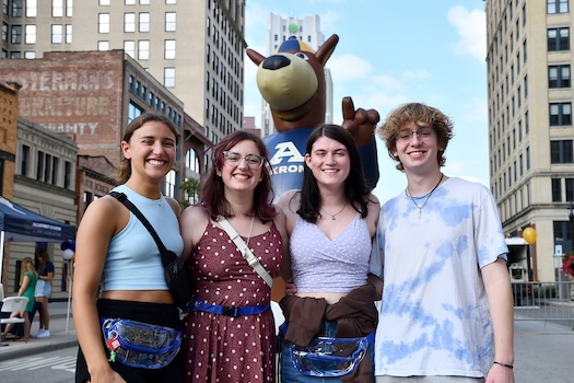 Four smiling young adults stand together on a city street with an inflatable kangaroo mascot and tall buildings in the background. It appears to be a sunny day with a festive atmosphere.