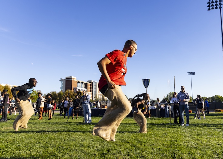 Students compete in a sack race on a grassy field during an outdoor campus event on a sunny day, with a crowd of spectators and university buildings visible in the background.