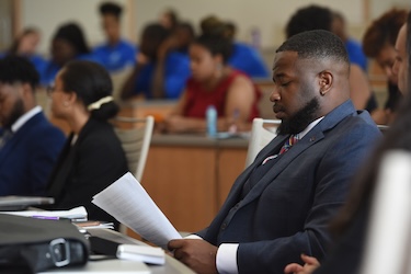 A man in a suit sits at a desk in a classroom, reading a document, with other students seated and listening attentively in the background.