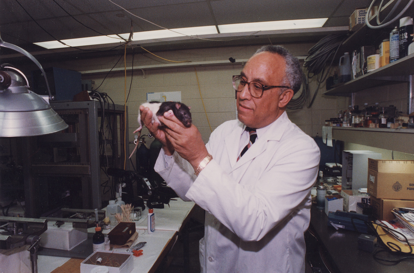 A scientist in a white lab coat holds a rat in a laboratory filled with equipment, shelves, and supplies, examining the animal under bright overhead lights.