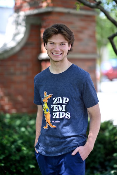 A young man with brown hair smiles at the camera, standing outdoors in front of a brick wall and greenery. He wears a blue t-shirt with a cartoon kangaroo and text that reads, ZAP EM ZIPS.