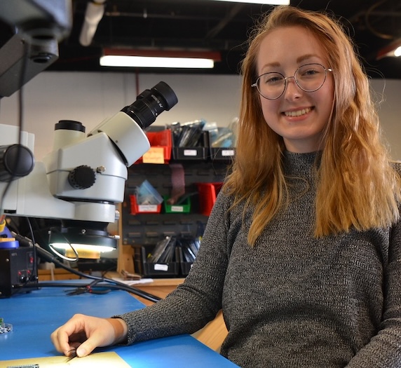 A woman with long red hair and glasses smiles while sitting at a workbench beside a microscope in a lab or workshop setting. Storage bins and various equipment are visible in the background.