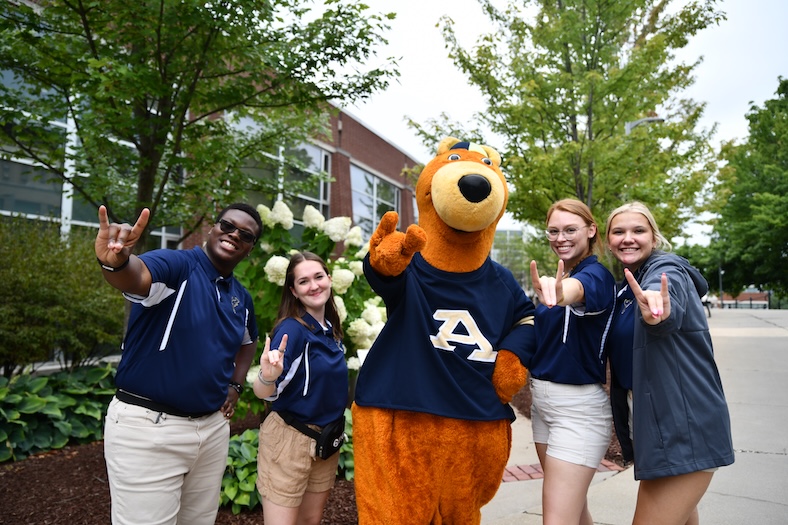 Four smiling students in navy University of Akron shirts pose with the Zippy kangaroo mascot outdoors, all making the zippy hand sign near green trees and a brick building.