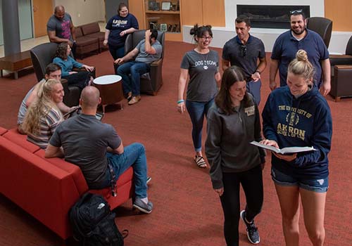 A group of college students socialize and interact in a lounge area with red couches and chairs. Some are seated and chatting, while others are standing or walking, holding papers and smiling.