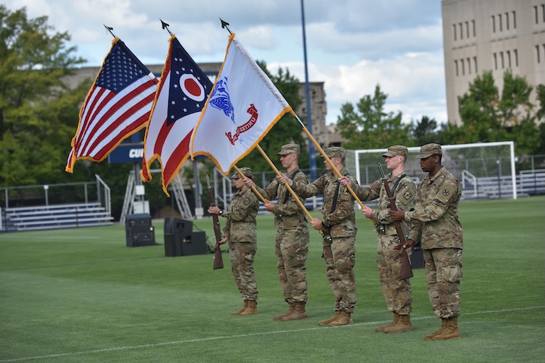 ROTC color guard stands at attention on a college athletic field, displaying the U.S. flag, Ohio state flag, and a unit flag, while armed cadets in camouflage uniforms stand at parade rest during a university convocation ceremony.