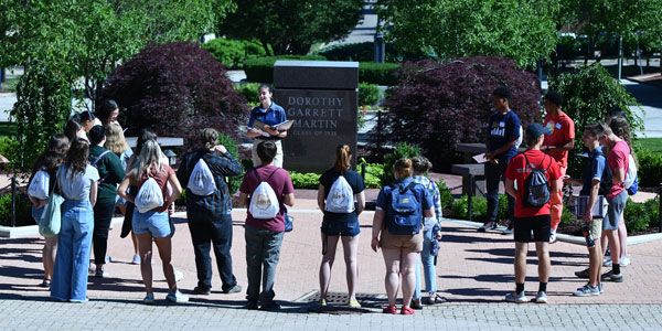 A campus tour guide addresses a group of prospective students and families gathered around the Dorothy Garrett Martin monument on a sunny day at the University of Akron.