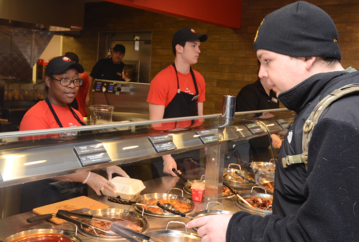 A customer in a black beanie and backpack is ordering food at a counter while two employees in red shirts and black aprons serve him from hot food trays. The setting appears to be a casual restaurant.