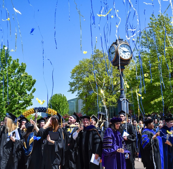 Graduates in caps and gowns celebrate outdoors as blue and yellow streamers fall. A clock tower and trees are in the background, with clear blue skies overhead.