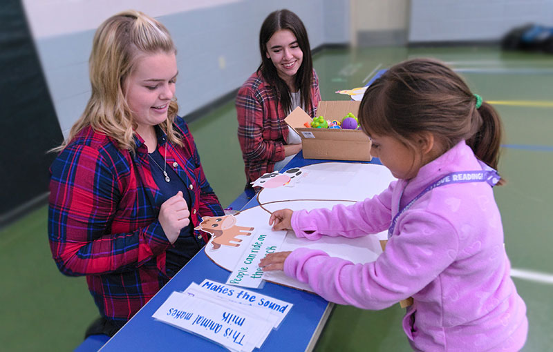 Two women sit at a table helping a young girl with an animal-themed learning activity involving cut-out animals and sentence strips in a classroom setting.