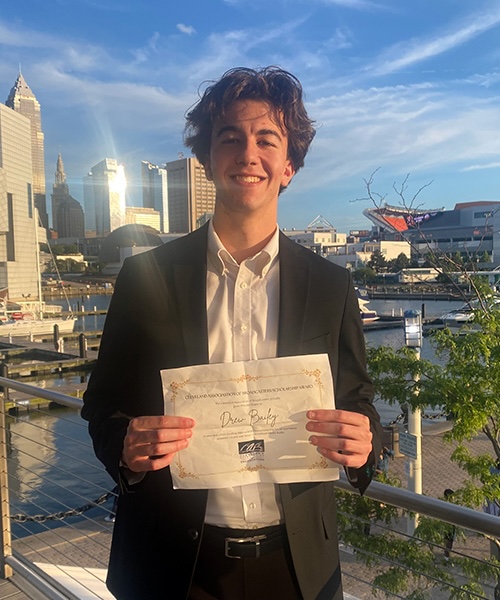 Gemini said An image of a young man with wavy brown hair, smiling and wearing a black suit jacket over a white collared shirt. He is standing outdoors on a waterfront walkway, holding a certificate from the Cleveland Association of Broadcasters with the name 