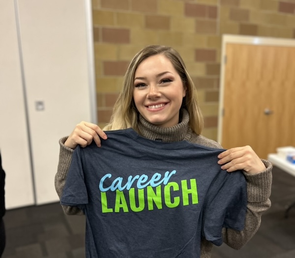 A smiling woman holds up a T-shirt that says Career Launch in blue and green lettering, standing indoors with beige brick walls and a wooden door in the background.