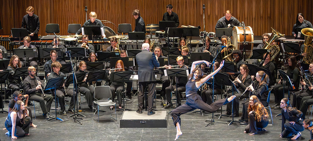 A concert band performs on stage, with musicians dressed in black seated in rows playing wind and percussion instruments. A conductor stands at the podium with their back to the camera. In the foreground, a dancer in a black outfit leaps gracefully mid-air, arms extended, while other dancers sit on the stage floor around her.