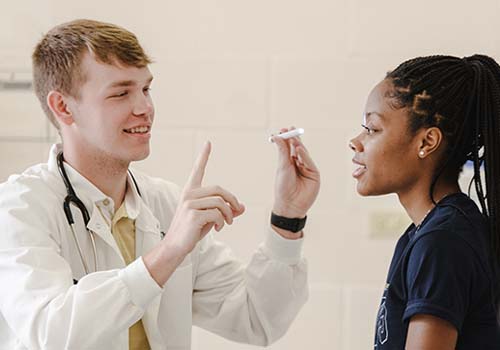 A doctor with a stethoscope performs an eye exam on a patient by holding up a light and raising a finger, while the patient looks forward and follows instructions.