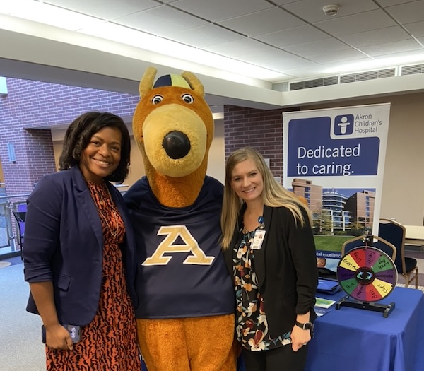 Two women stand next to a kangaroo mascot wearing an A shirt at an Akron Children’s Hospital event. A table with a colorful prize wheel and a hospital sign reading Dedicated to caring. is in the background.