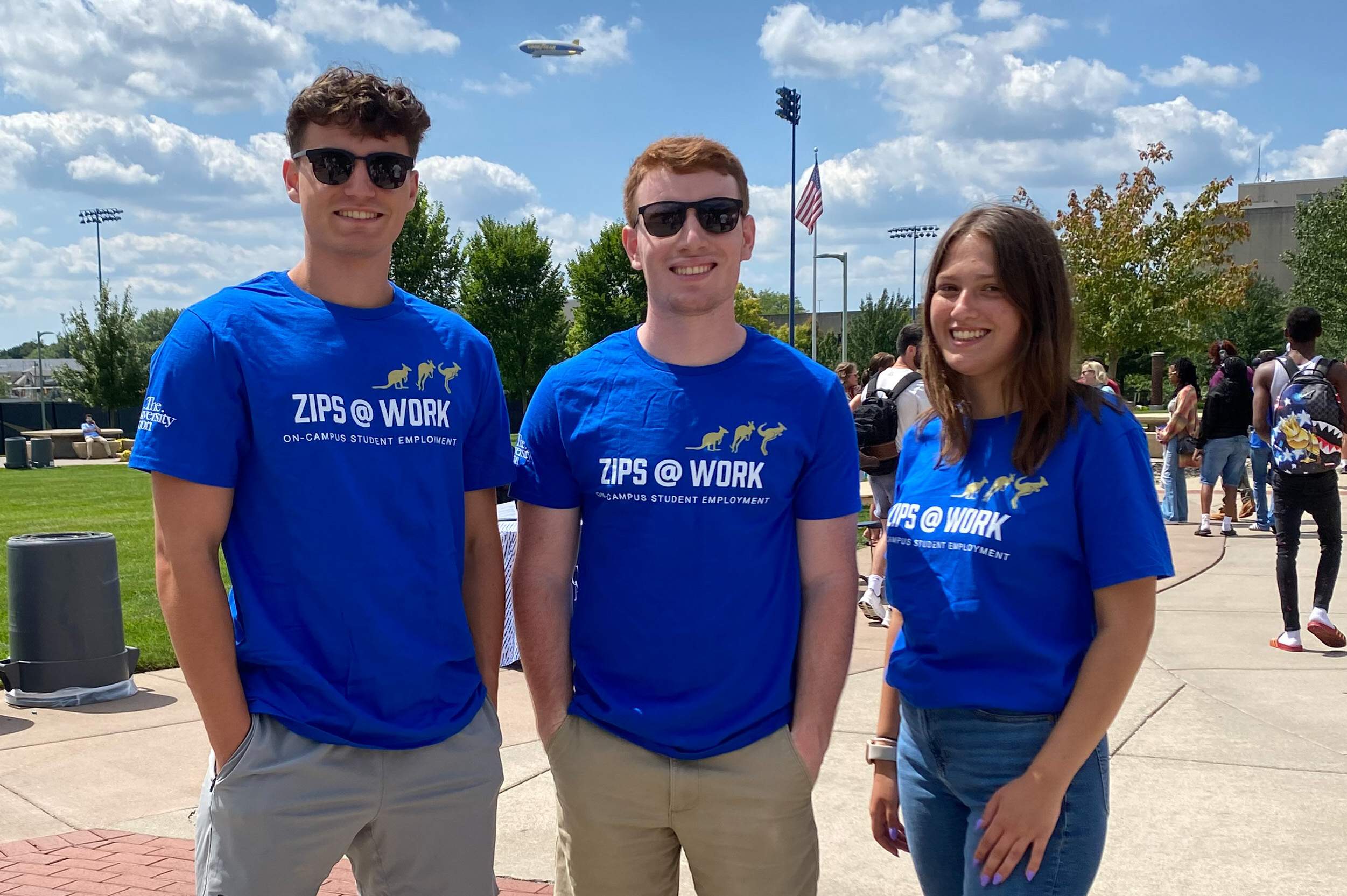 Three students standing outside on Coleman Common at UA wearing Zips@Work t-shirts and smiling