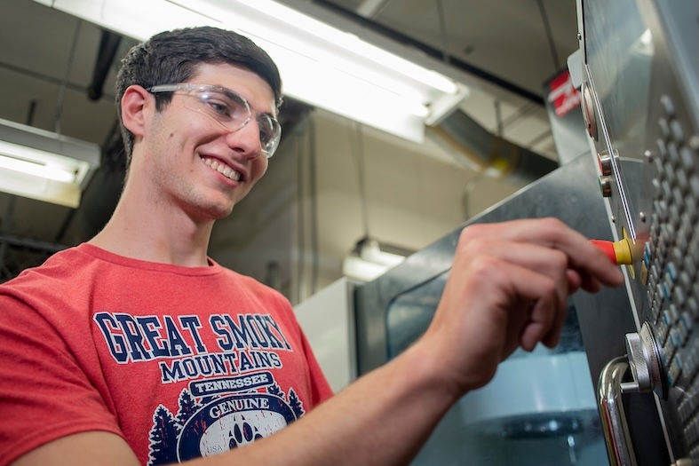 A young man wearing safety goggles and a red Great Smoky Mountains T-shirt operates machinery, pressing a button on a control panel in an industrial or workshop setting.