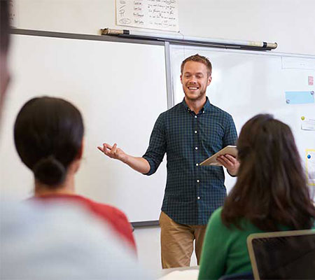 Instructor speaks at the front of a classroom while holding notes and leading a discussion.