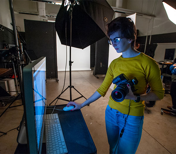 Student works at a computer while holding a camera in a photography studio.
