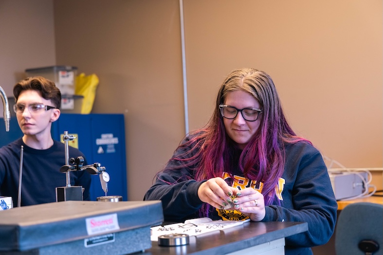 A student with purple hair and glasses works with small components at a lab bench, while another student wearing safety goggles observes in the background of a college engineering or manufacturing lab.