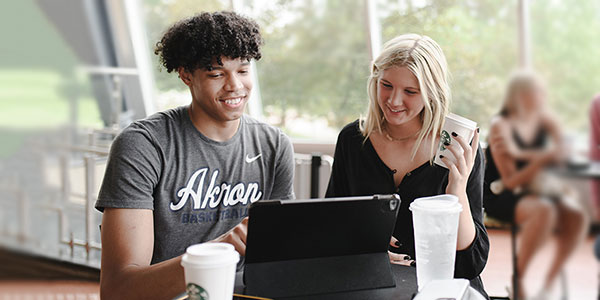 Two University of Akron students smile while looking at a tablet together at a café table with Starbucks drinks, in a bright, windowed campus building.