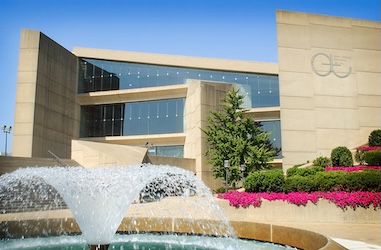 A modern beige concrete building with large glass windows, a fountain in the foreground, colorful flowers, and a clear blue sky. A stylized QE logo is visible on the buildings facade.