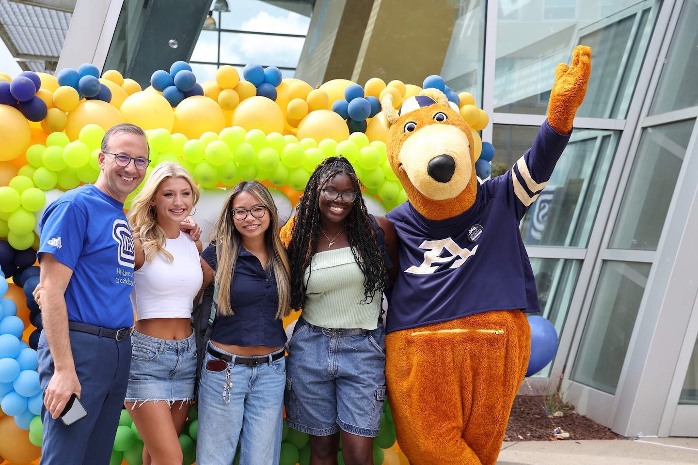 Three students and a staff member pose with Zippy, the University of Akron's kangaroo mascot, in front of a colorful balloon arch at a campus event.