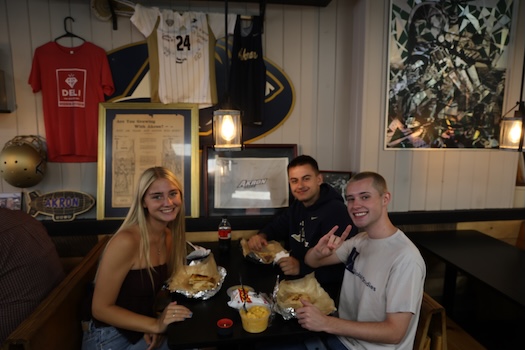Three young adults sit at a restaurant table, smiling at the camera with food and drinks in front of them. The wall behind them is decorated with sports jerseys, framed pictures, and memorabilia.
