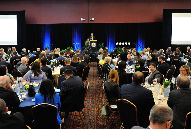 A large group of people seated at round tables during a formal banquet or conference, facing a speaker at a podium on a stage with black curtains and blue lighting.